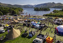 Outdoor festival with tents, stages, and crowds near a lake surrounded by mountains.