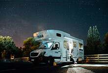 Motorhome parked under a starry night sky with trees and a wooden fence nearby.