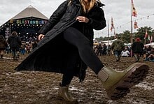 Woman in wellington boots at outdoor festival with muddy ground and stage in background.